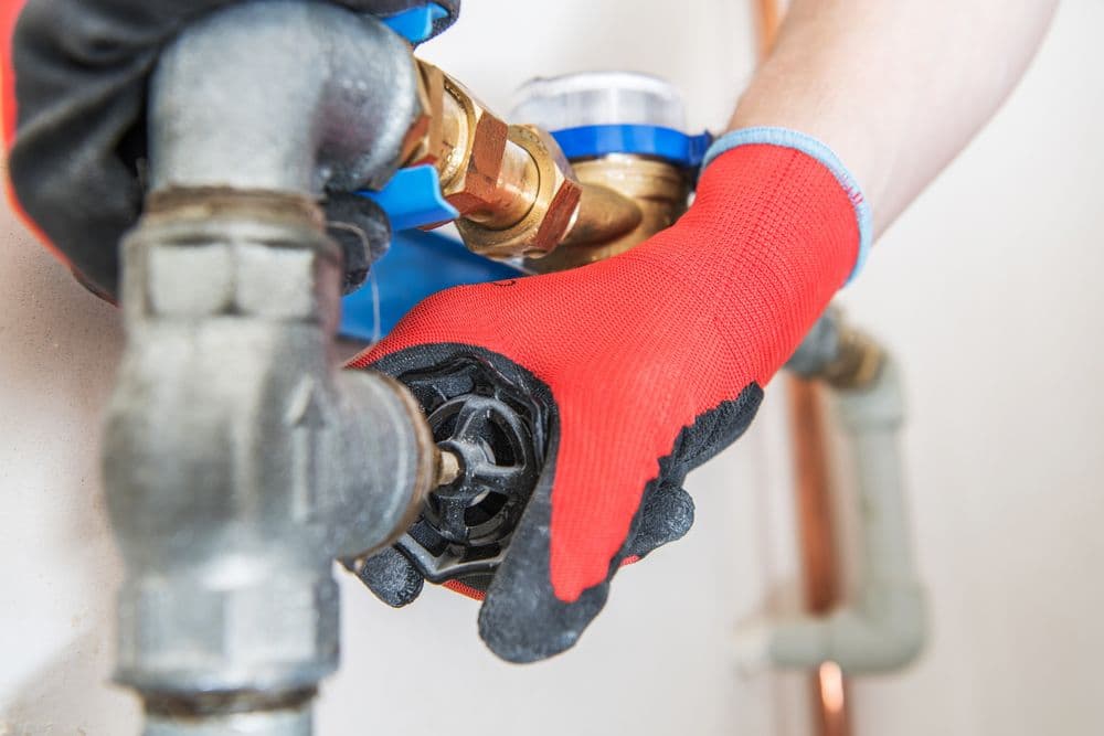 Person in red gloves adjusting a water valve on plumbing pipes indoors.